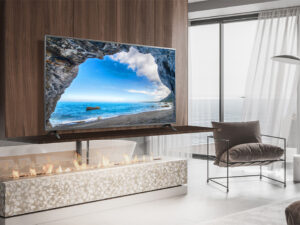 In a simple bedroom overlooking the sea, there is a TV on a wall shelf. The blue sea scenery appears bright and clear on the TV screen.